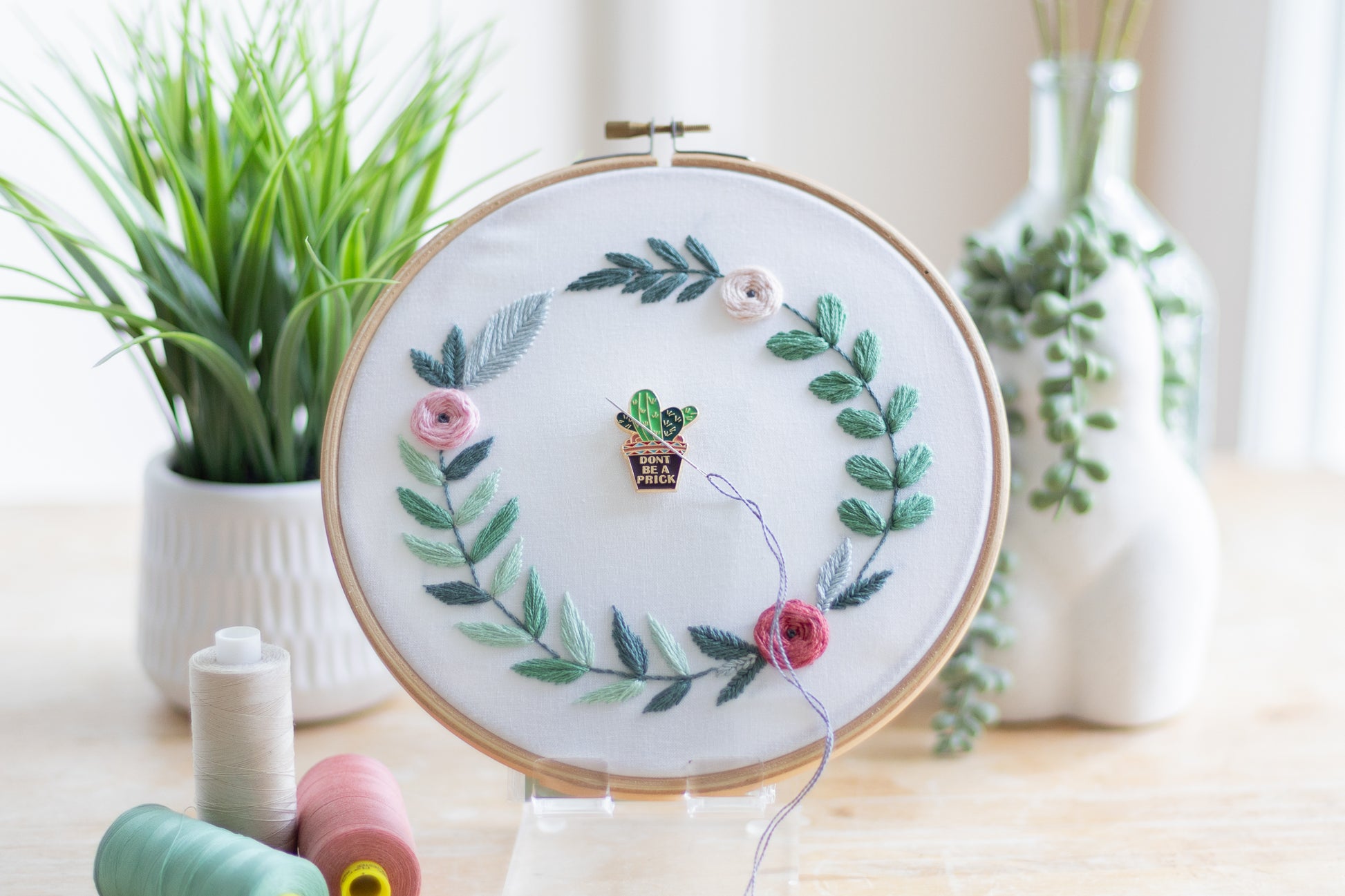 Focal Point: Cute potted cactus with the words "Don't be a prick" on the pot. There is a sewing needle on the cactus which is a magnetic needle minder. The needle minder is secured with a magnet to an embroidery hoop with floral and leaf design on a wooden table with plants in the background is
